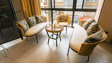 A modern lounge area with wicker furniture, a glass coffee table, and large windows offering a city view.