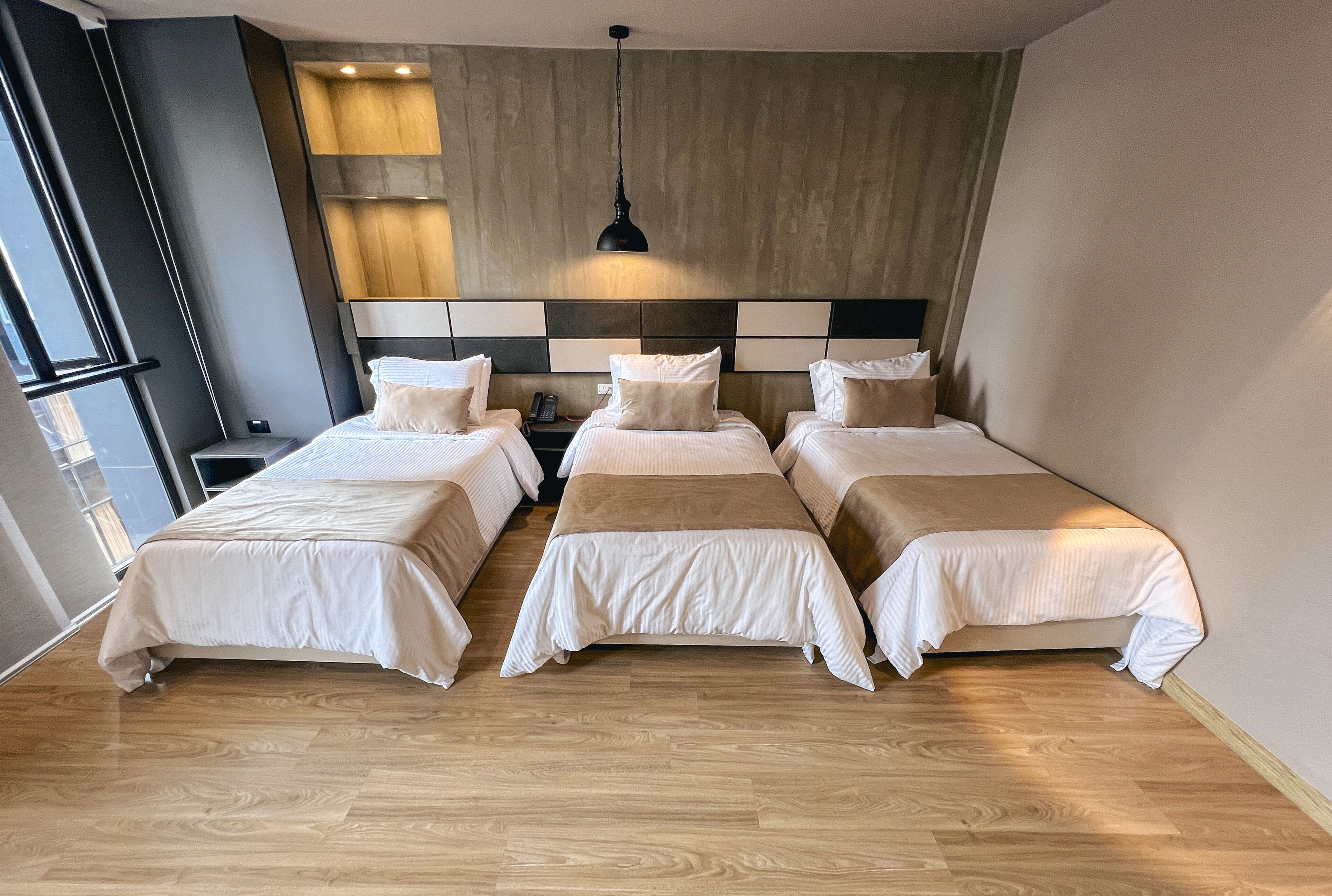 A hotel room with three single beds, a wooden floor, a large window, and a modern ceiling light.