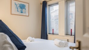 A neatly made bed with rolled towels, a framed artwork on the wall, and a window with blinds. - Central Apartment in Cathedral City of Ripon (RIPON)