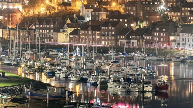 A marina with boats docked, a castle illuminated in the background, and a town with buildings along the waterfront. - Central Apartment in Cathedral City of Ripon (RIPON)