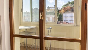 Ein Balkon mit Metallstühlen und einem Tisch, eine Holztür und Blick auf Gebäude und Bäume durch ein Fenster.