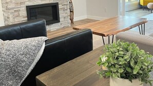 A modern living room with a stone fireplace, a flat-screen TV mounted above it, a dining table with chairs, and a yellow sofa in the background.