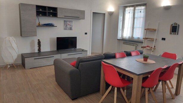 Een moderne leefruimte met een grijze bank, een eettafel met rode stoelen, een televisie op een grijze dressoir, en een raam met gordijnen.