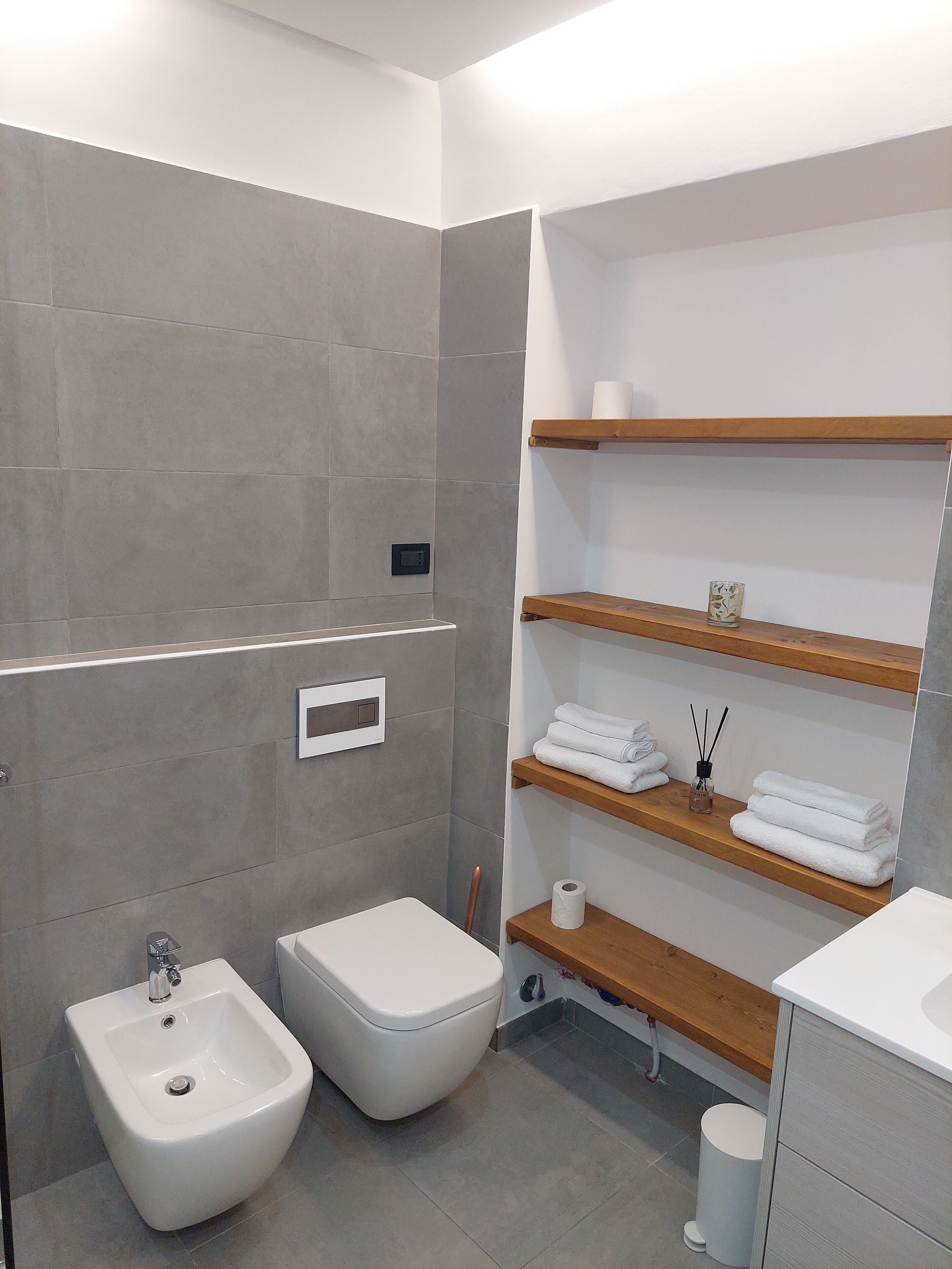 Een moderne badkamer met grijze betegelde wanden, een bidet, toilet en houten planken met handdoeken en toiletartikelen.
