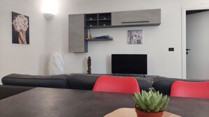 Een moderne woonkamer met een donkerbruine tafel, rode stoelen, een grijze bank, een flatscreen televisie, een grijze boekenkast en een pot met een vetplant.