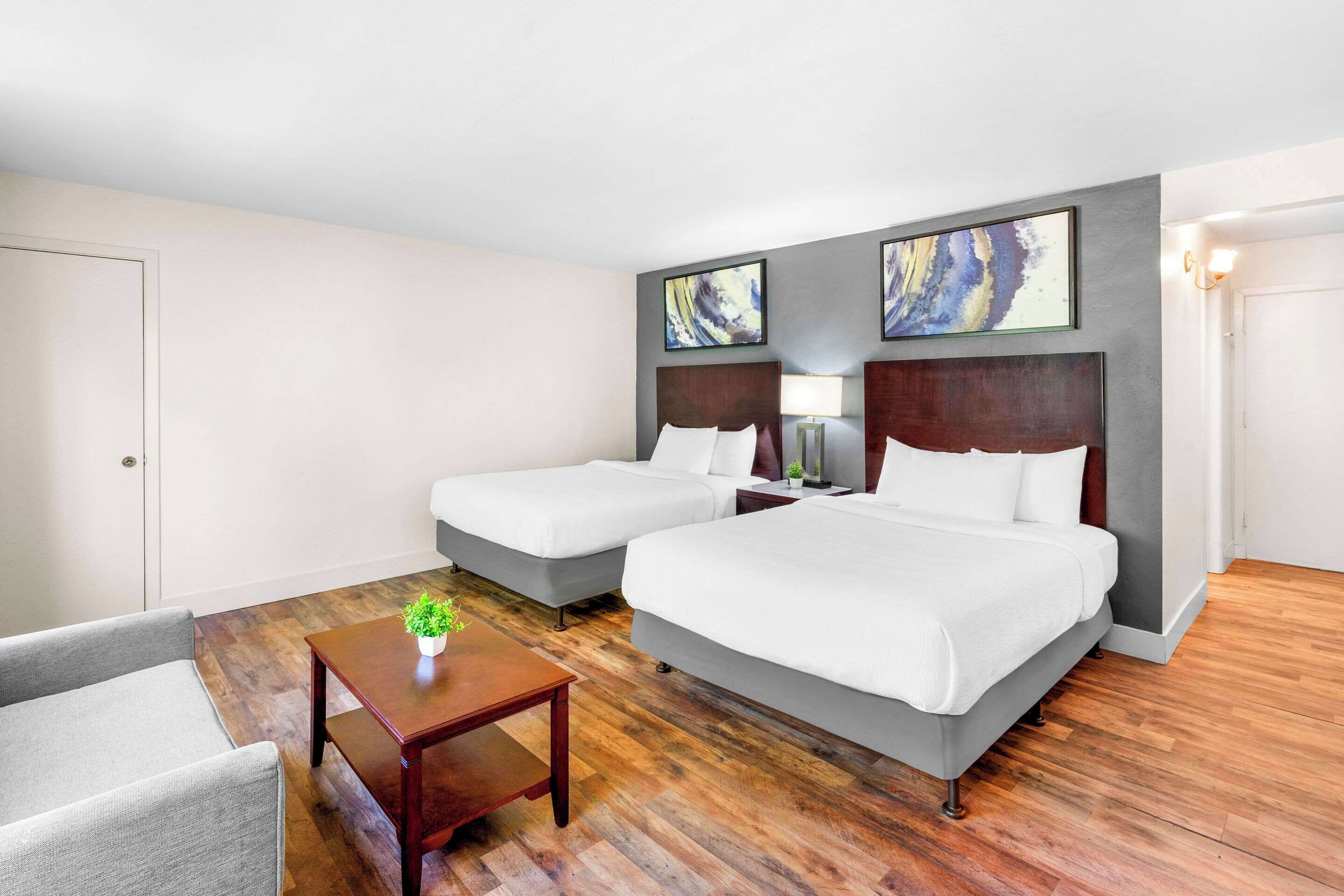 Quarto de hotel com duas camas, uma mesinha com uma planta e um sofá cinzento.