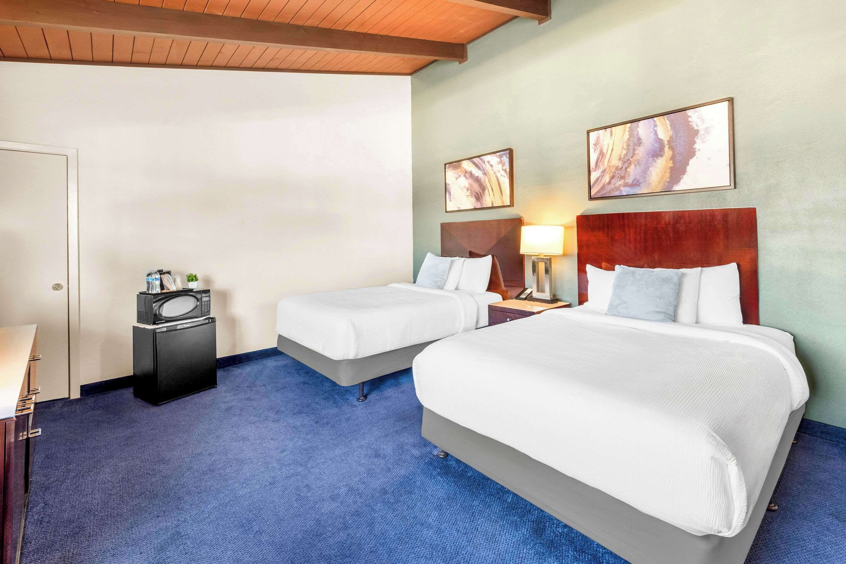 A hotel room with two beds, a desk, a microwave, and artwork on the walls.