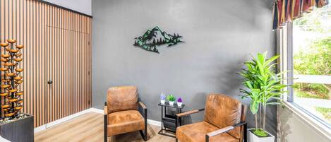 Una habitación moderna con dos sillones de cuero, piso de madera, un adorno en la pared, una pequeña mesa con plantas y un ventanal con vistas al verdor.
