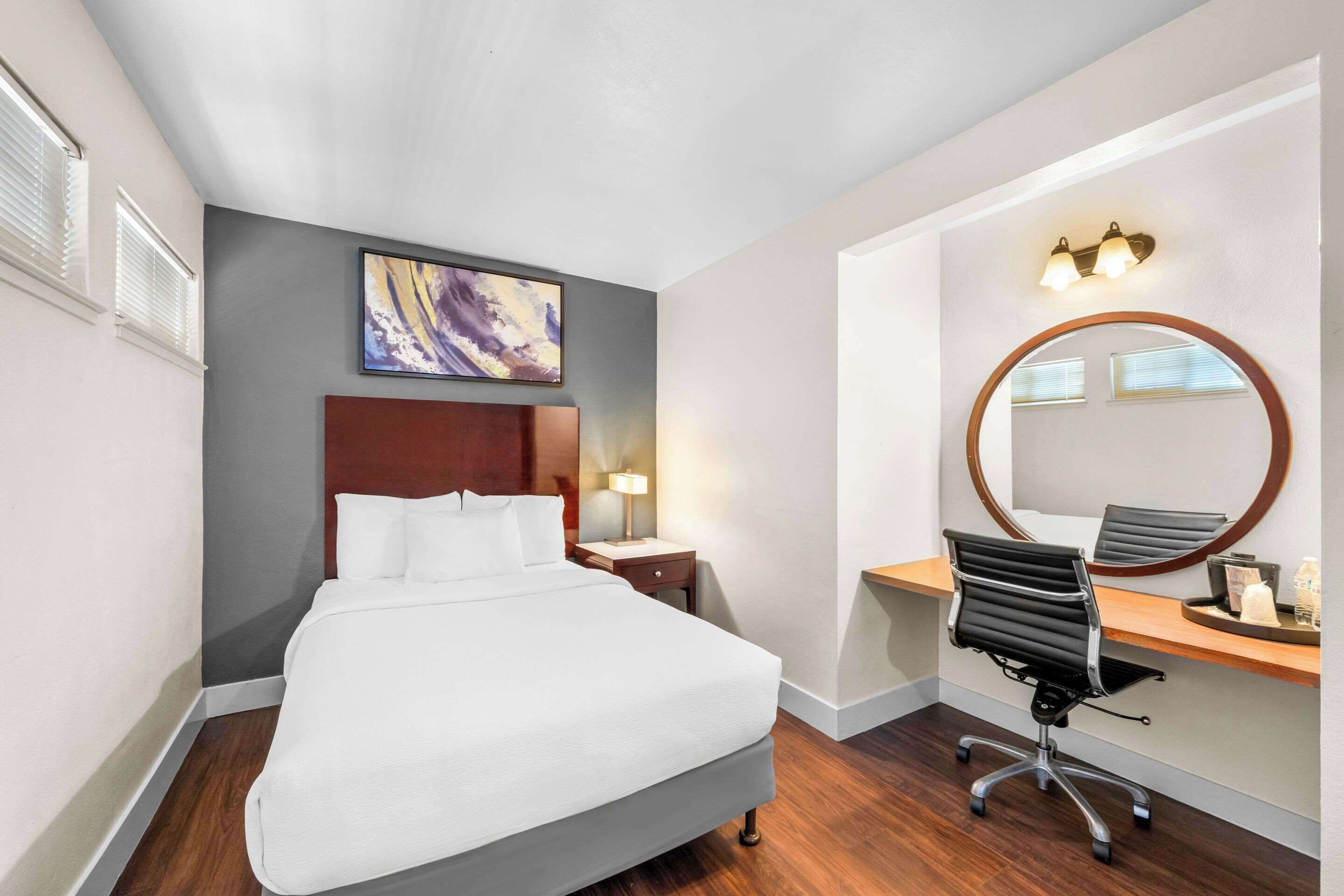 A hotel room with a bed, a desk with a chair, a round mirror, and a painting on the wall.