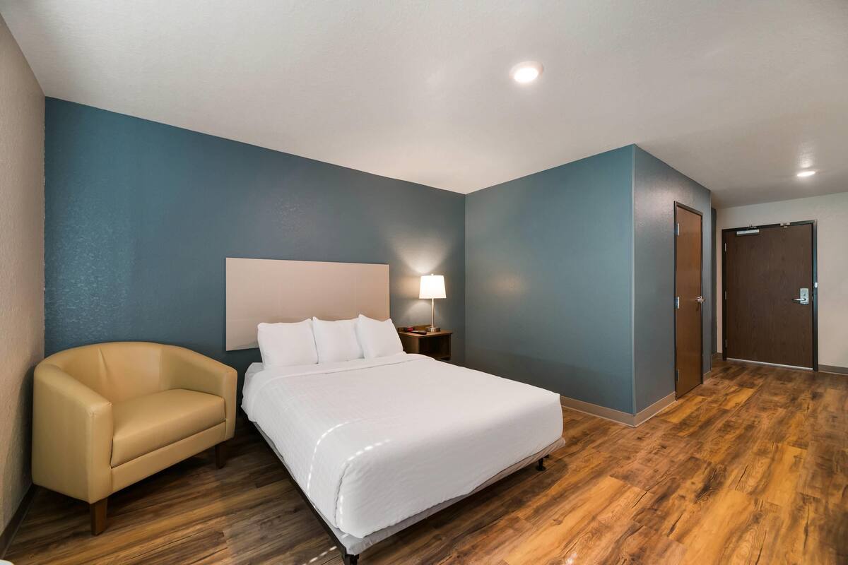 A hotel room with a bed, a chair, a nightstand with a lamp, and a wooden floor.