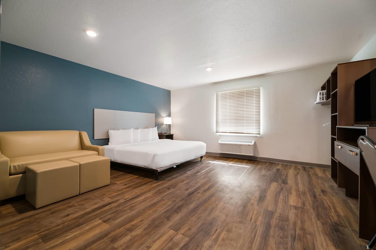 A modern hotel room with a bed, a sofa, a desk, and a window with blinds.