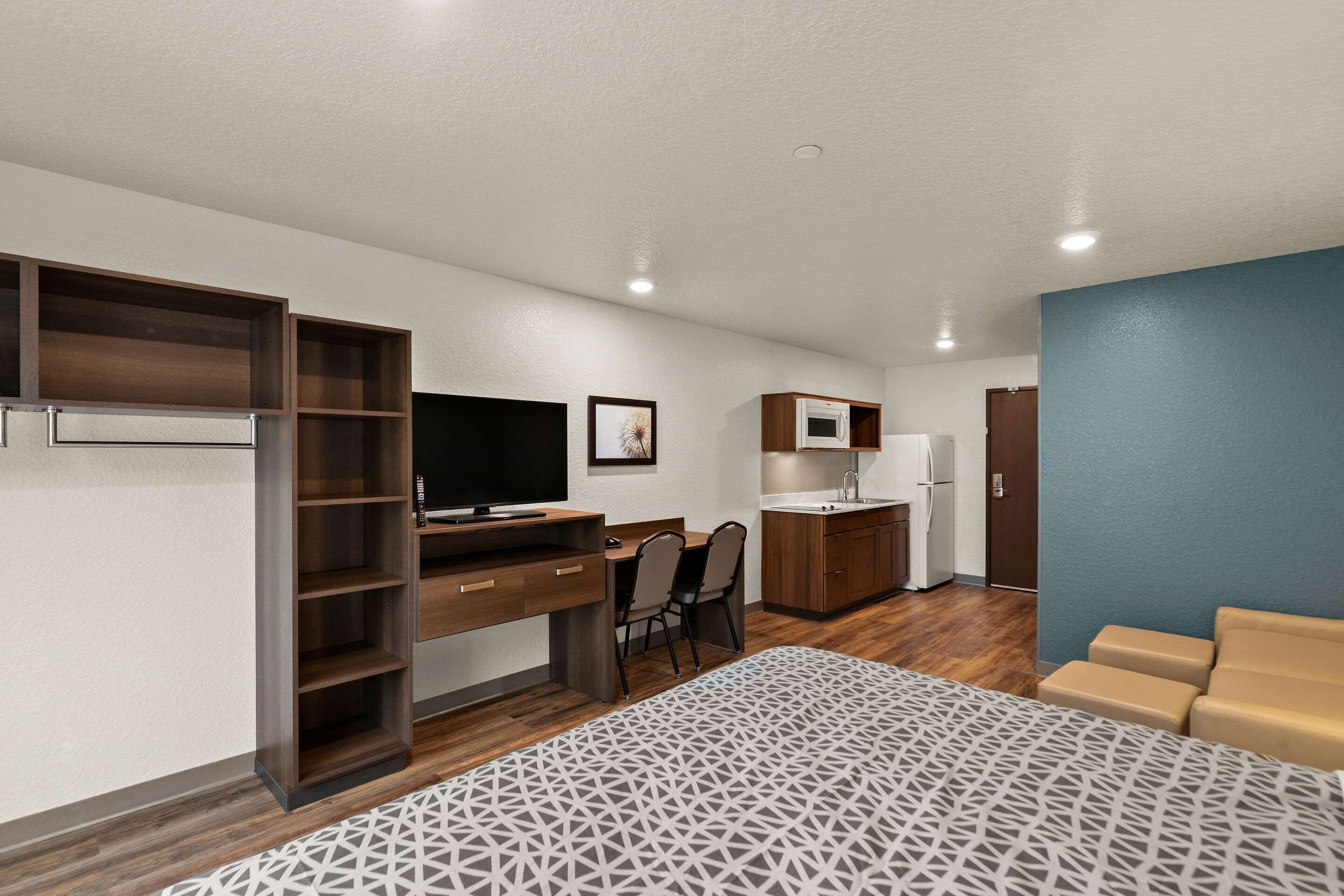 A compact hotel room with a bed, a desk, a kitchenette, and a seating area.