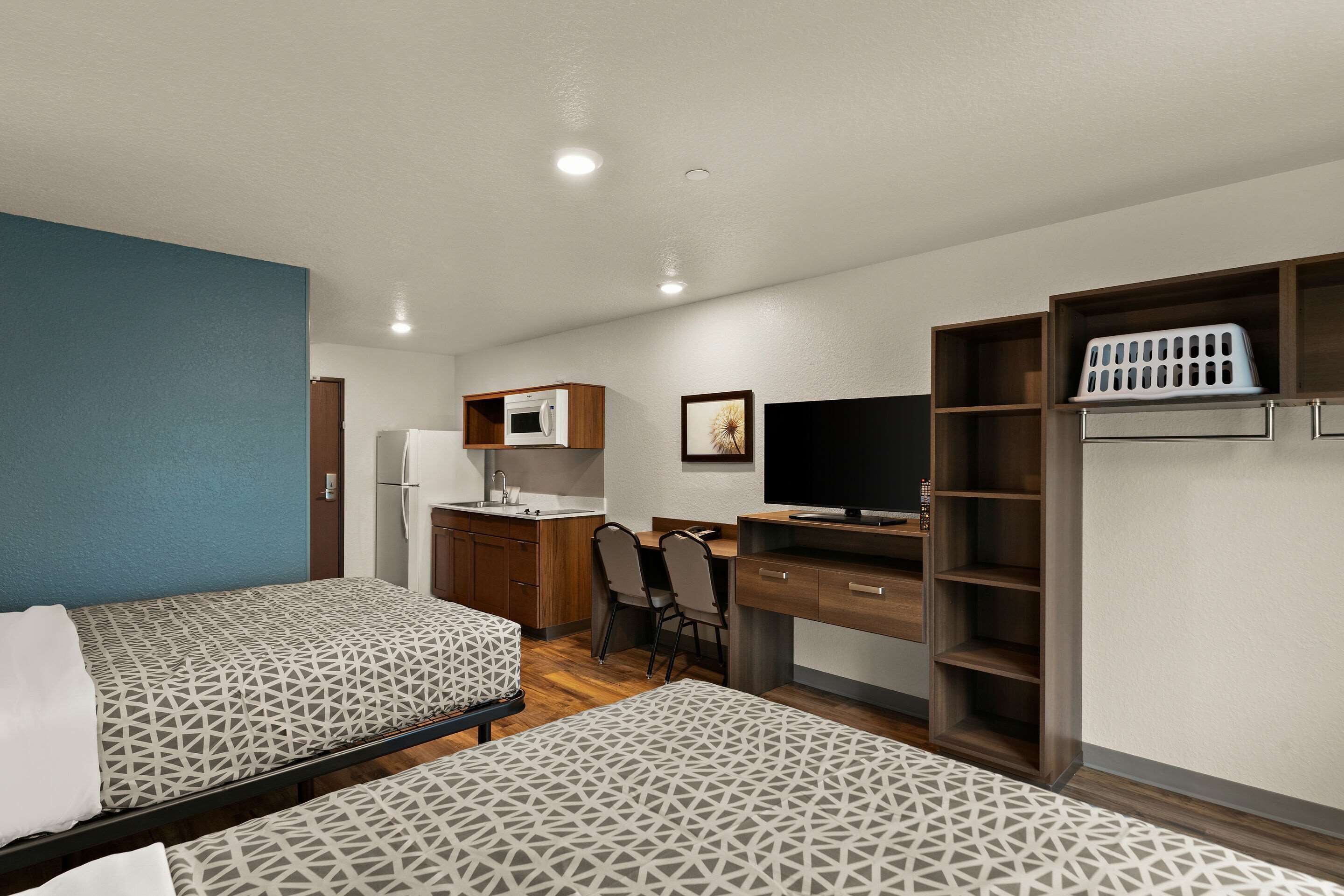Habitación de hotel con dos camas, kitchenette, escritorio con silla, televisor y armario.