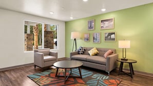 Un salon moderne avec un canapé, deux fauteuils, une table basse et un tapis. On y trouve deux lampadaires et une fenêtre donnant sur des arbres et un immeuble.