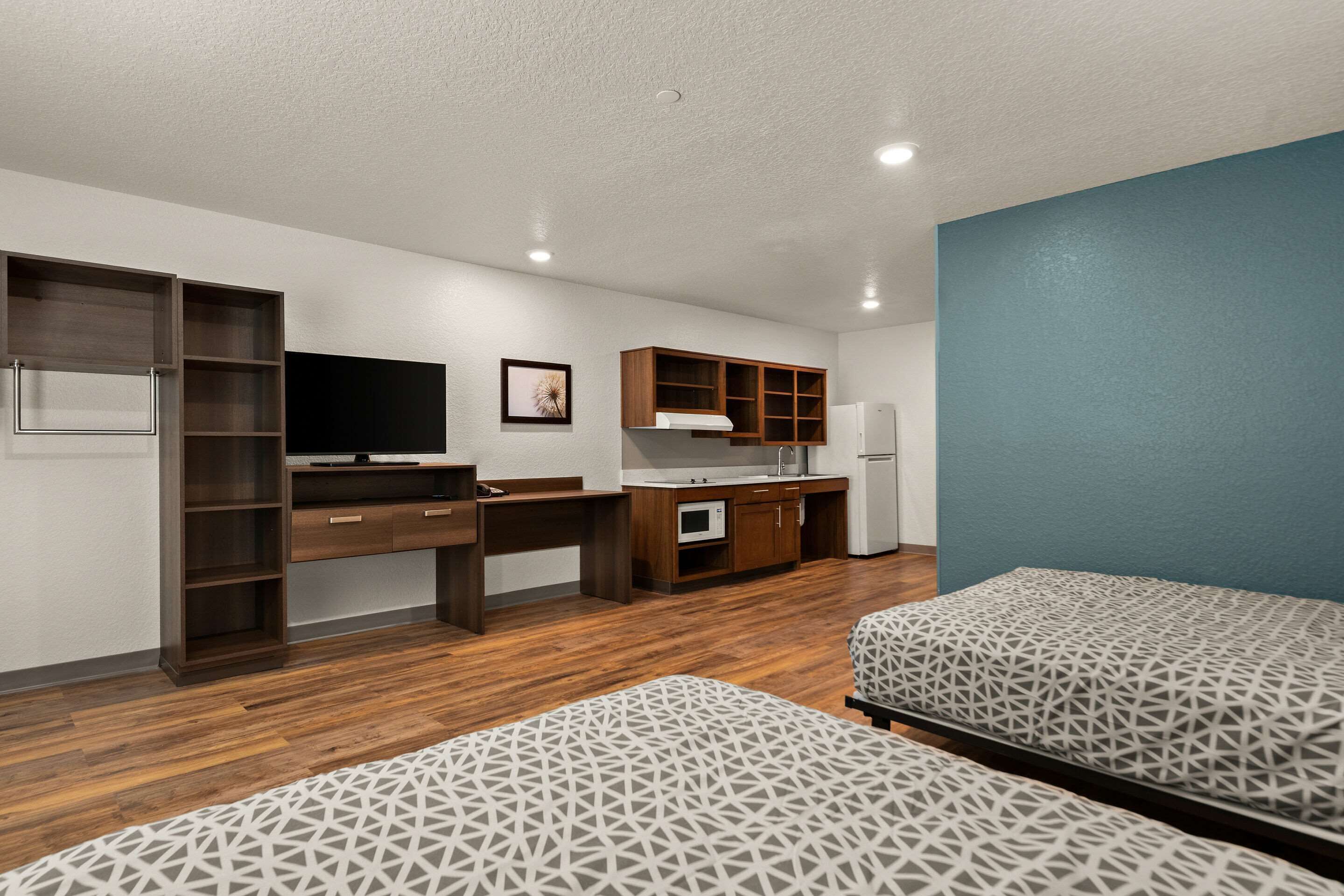 A modern hotel room with a kitchenette, two beds, a desk, and a TV.