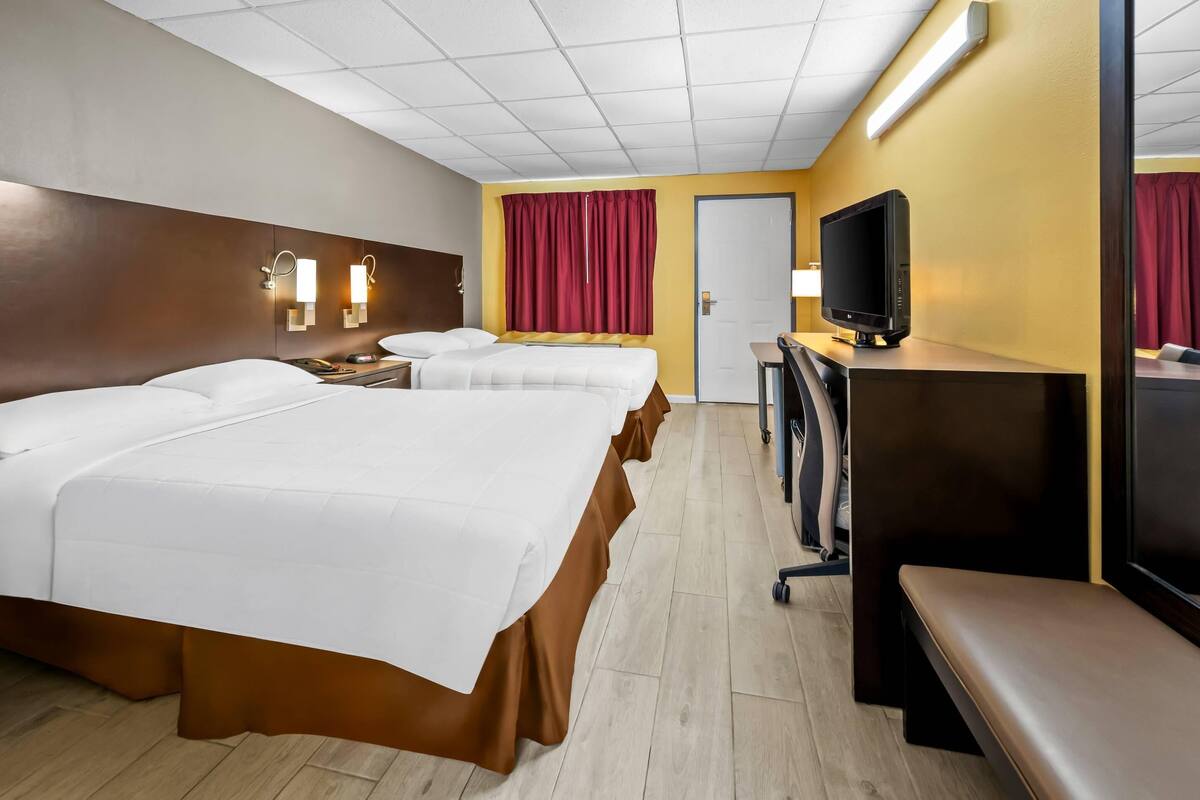 Quarto de hotel com duas camas, uma mesa de trabalho, uma televisão, um banco e uma porta.