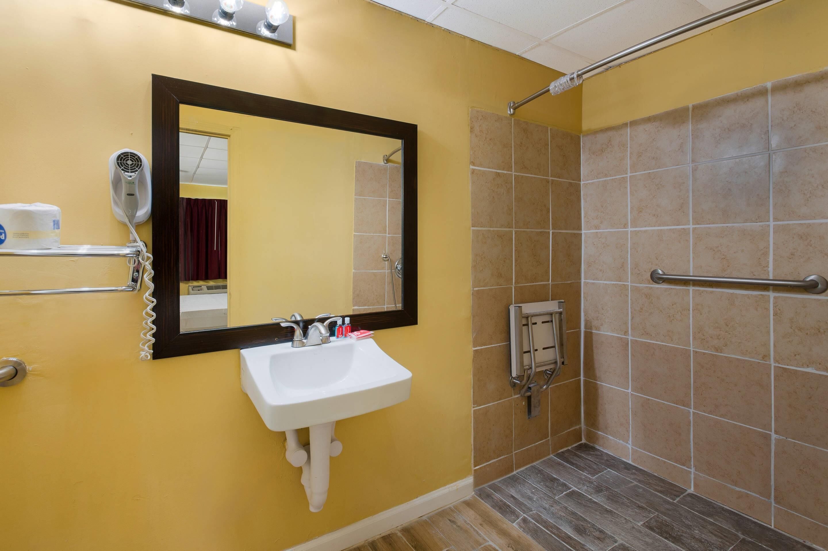 A bathroom with a yellow wall, a white sink, a mirror, a shower with a grab bar, and a hairdryer on a shelf.