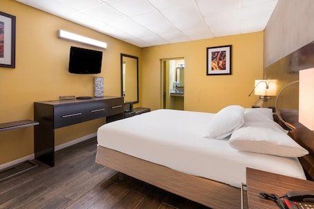 A hotel room with a bed, a desk with a telephone, a mirror, and a television mounted on the wall.