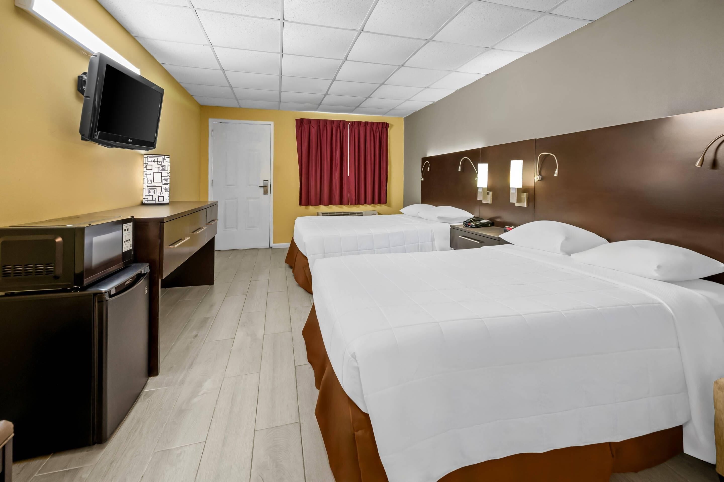 A hotel room with a flat-screen TV, a minibar, two beds with white bedding, a wooden headboard, and a red curtain.