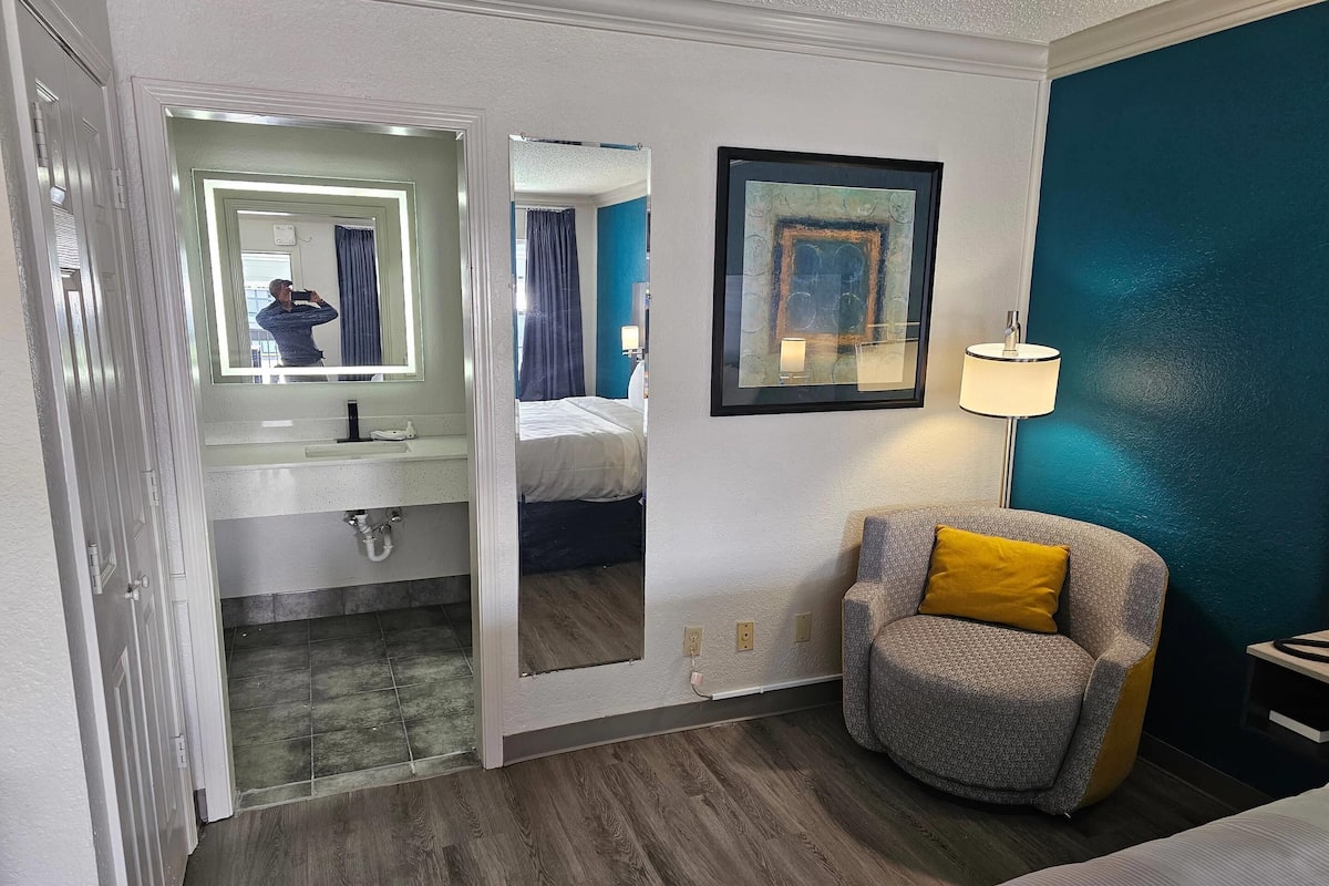 A hotel room with a bathroom, a bed, a chair, and a painting on the wall.