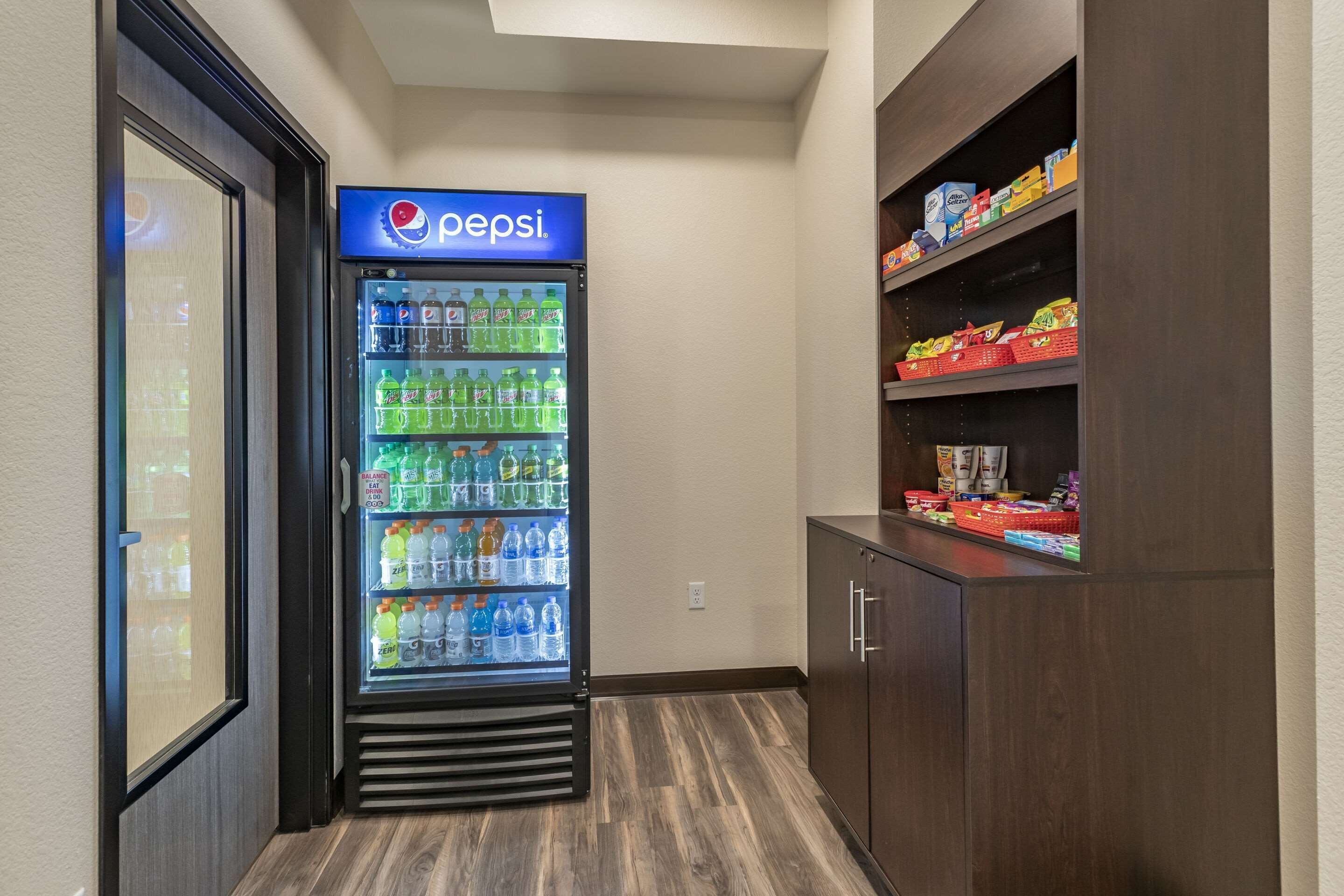 A hotel room with a Pepsi refrigerator stocked with various beverages, a wooden cabinet with snacks, and a door with a view of the outside.