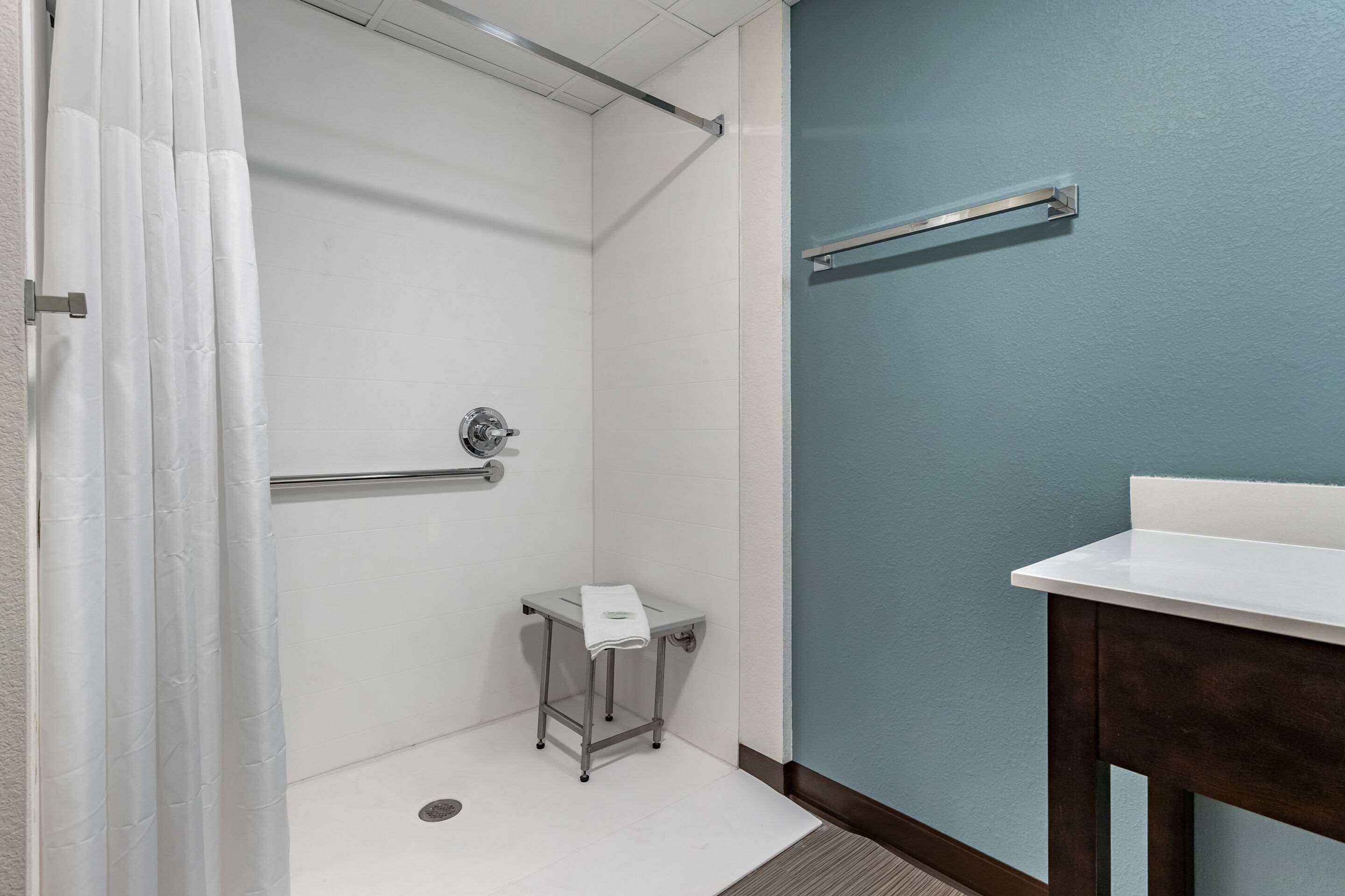 A bathroom with a shower, a small table with a towel, a handrail, and a sink.