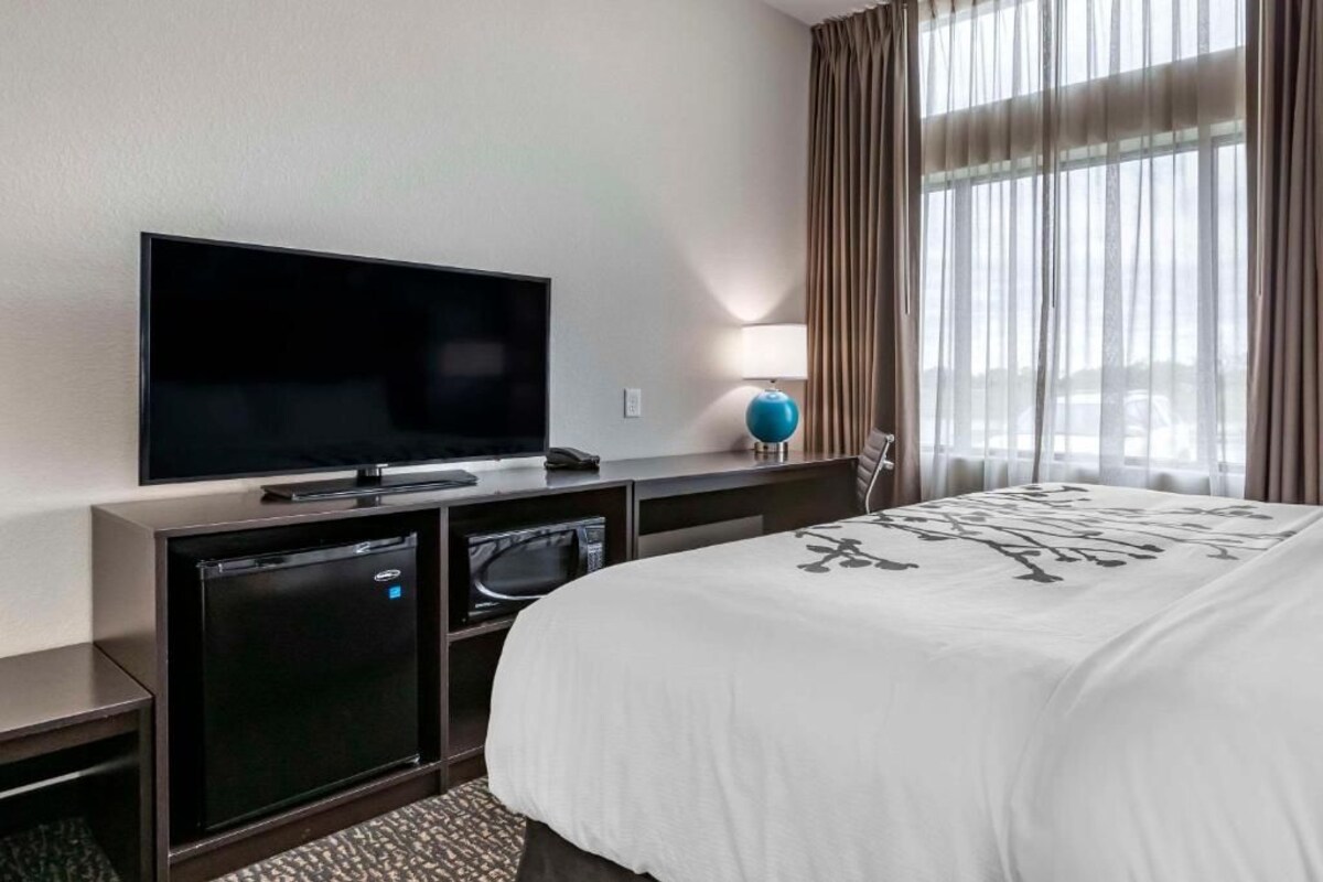 A hotel room with a flat-screen TV, a mini-fridge, a microwave, a bed with a white bedspread, a lamp, and a window with curtains.