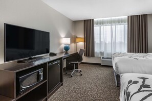 A hotel room with a flat-screen TV, a desk with a lamp, a chair, a bed, and a large window with curtains.