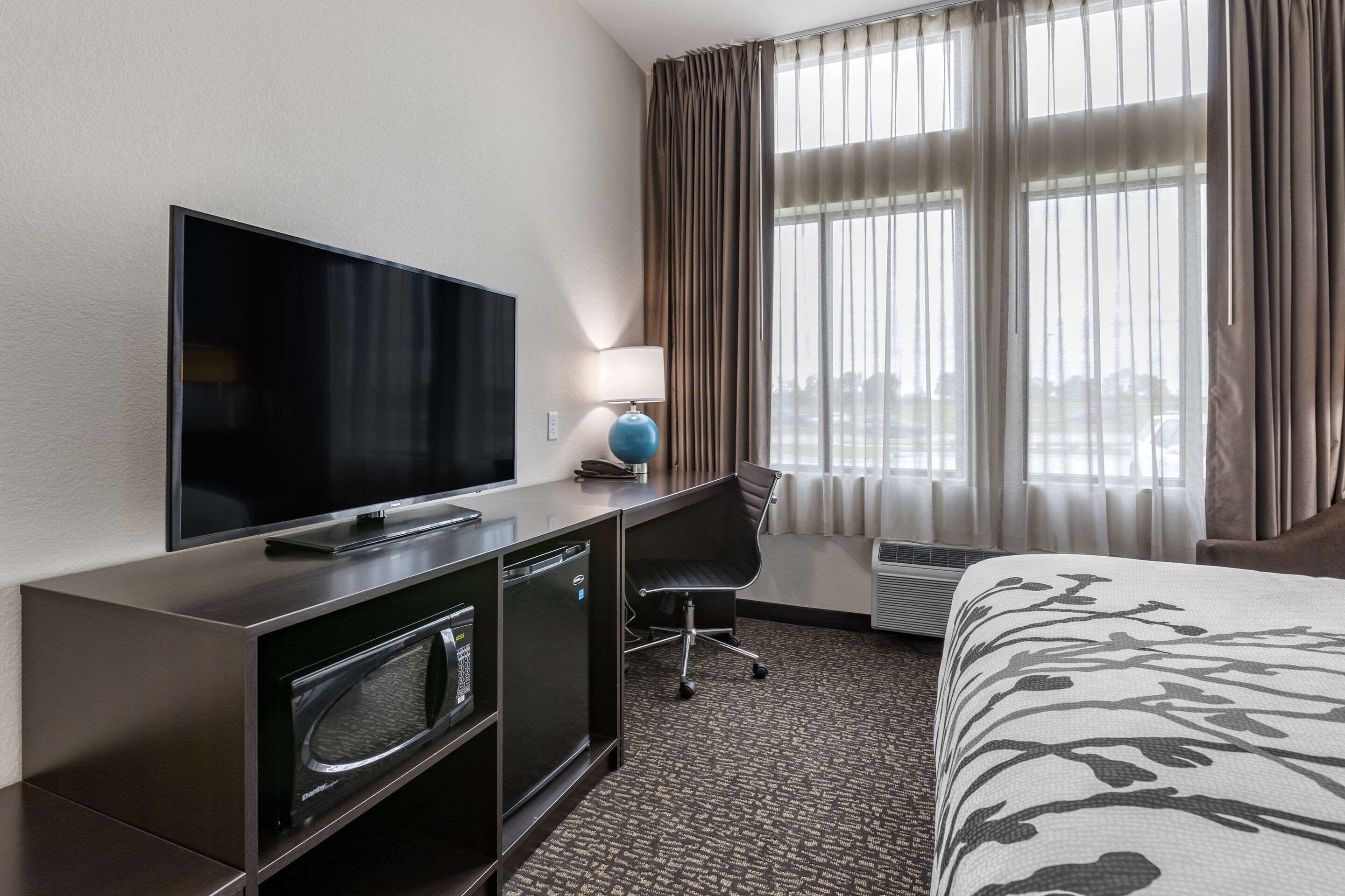 A hotel room with a flat-screen TV, a desk with a lamp, a chair, and a bed with a patterned bedspread.