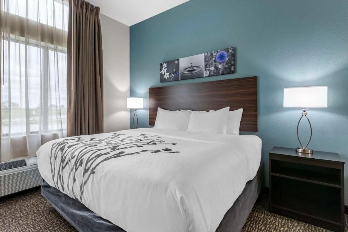 A hotel room with a large bed, two bedside lamps, a wall-mounted artwork, and a window with curtains.
