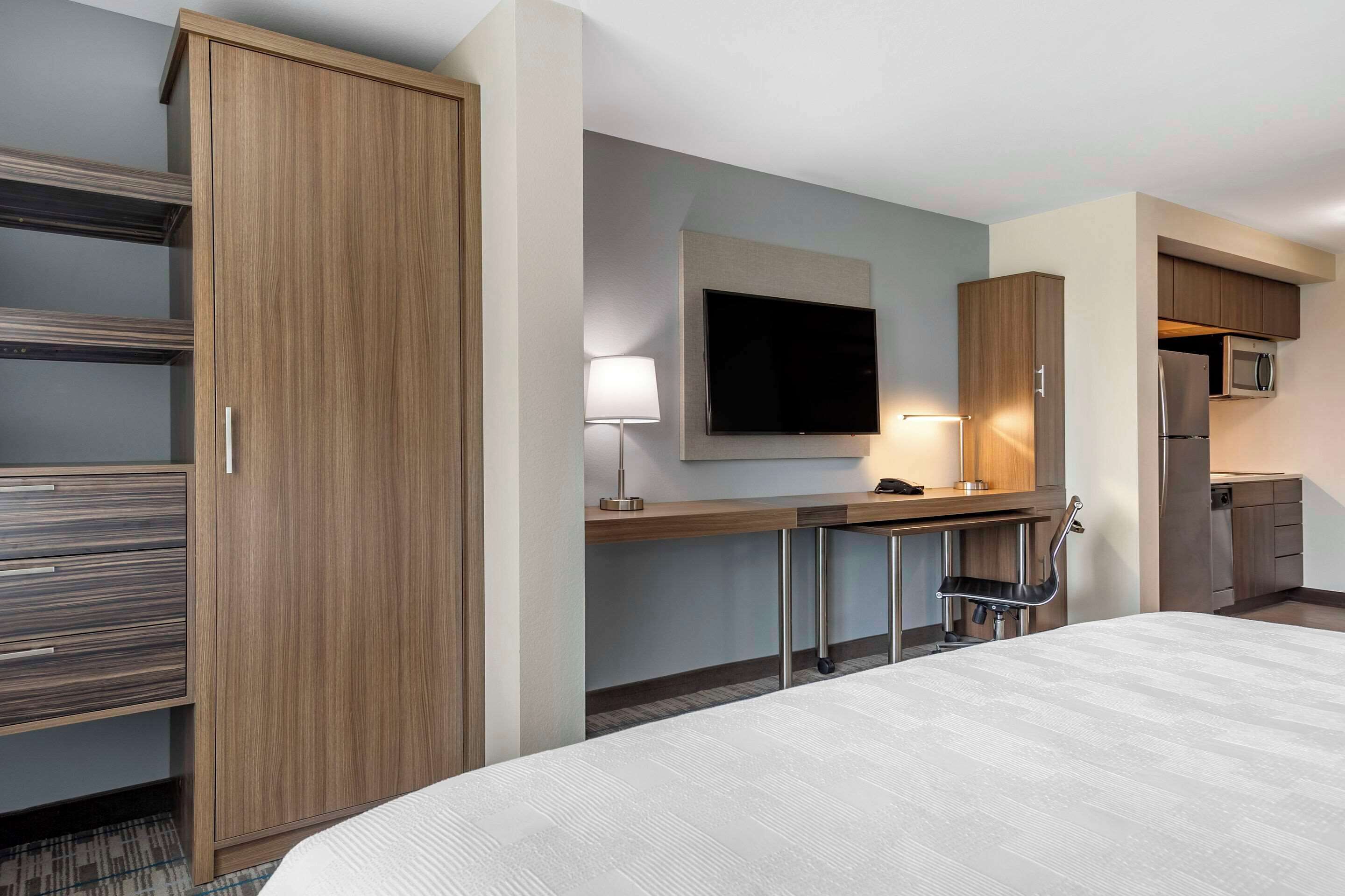 A hotel room with a large bed, a TV mounted on the wall, a desk with a lamp, and a kitchenette with a microwave and refrigerator.