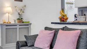 A living room with a grey sofa, pink pillows, a small table with a lamp, and a fruit basket.