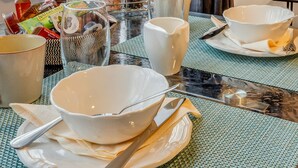 A breakfast table set with bowls, plates, cutlery, and a coffee maker, with a view of buildings through sheer curtains.