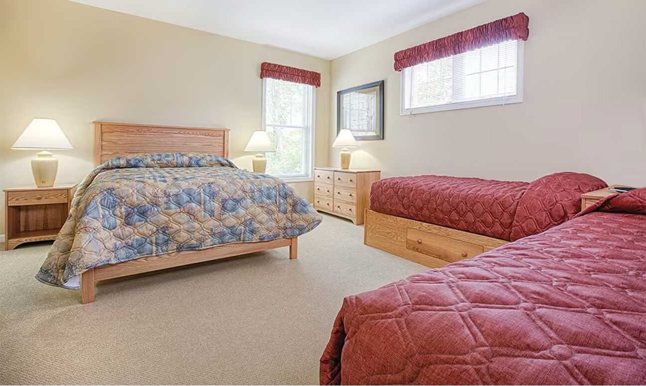 Une chambre à coucher avec un grand lit, un plus petit lit, deux tables de chevet avec des lampes, une commode et deux fenêtres avec des rideaux.