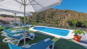 Une piscine entourée de chaises longues et de parasols, elle-même bordée d’un jardin aménagé et d’un paysage vallonné en arrière-plan.