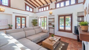 Un salón espacioso con un amplio sofá seccional color beige, una mesa de centro de madera y un televisor sobre un mueble de madera. Varias ventanas ofrecen luz natural y vistas al exterior.