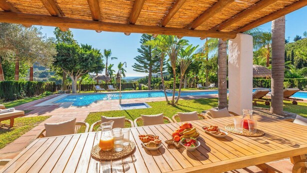 Una mesa de madera con alimentos para el desayuno y vista a una piscina y paisaje tropical.
