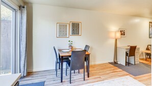 Un coin repas avec une table, des chaises et une lampe. Il y a une fenêtre avec des rideaux, une plante sur la table et des tableaux encadrés au mur.