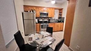 Cocina compacta con armarios de madera, un refrigerador de acero inoxidable, un microondas y una estufa. Hay una mesa de comedor con un mantel individual en el centro de la habitación.