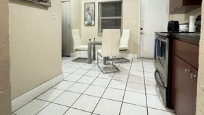 Una cocina con suelo de baldosas blancas, zona de comedor con mesa y sillas, y un refrigerador.