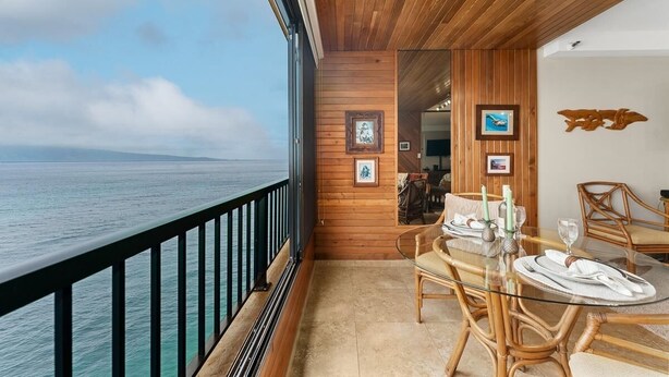 A balcony with a wooden railing overlooking the sea, a dining table set with white tableware, and framed pictures on the wall.