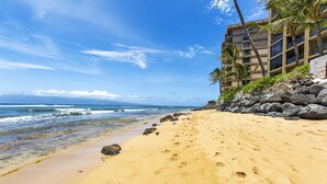 A beach with a clear view of the ocean, a building with a rocky shoreline, and palm trees.