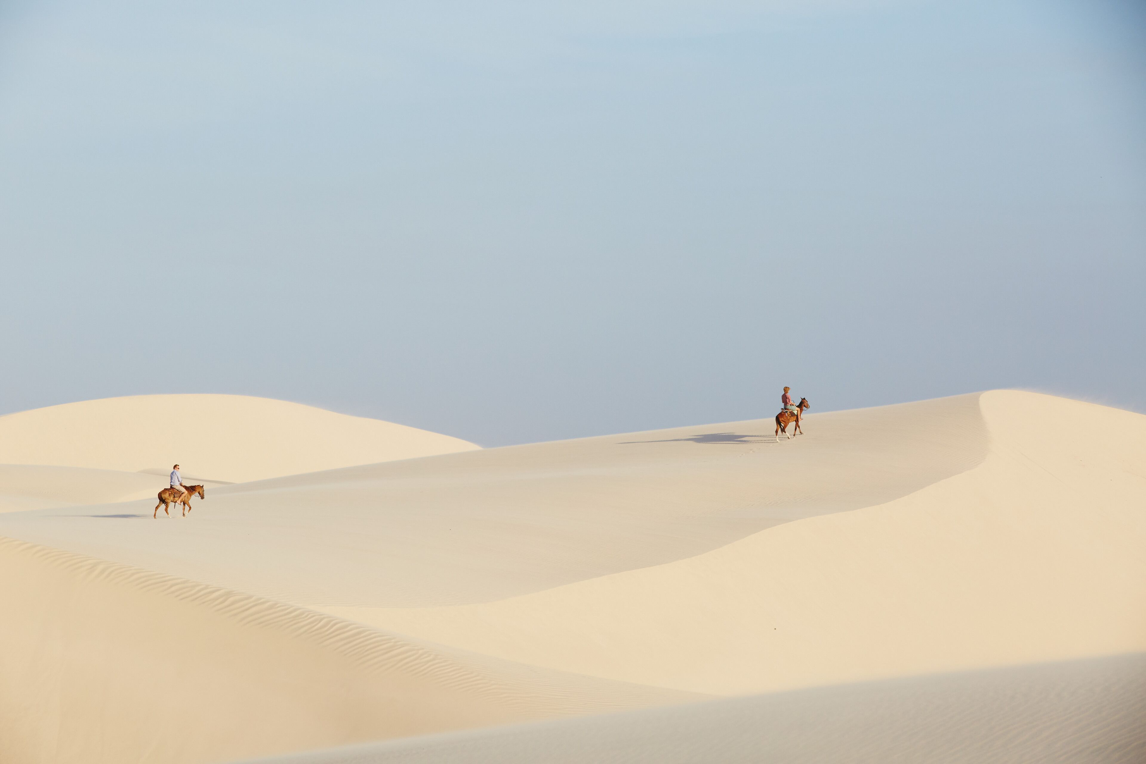 A desert landscape with sand dunes and two people riding horses.