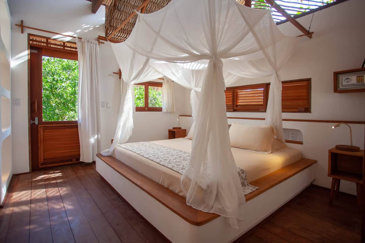 Un dormitorio con suelo de madera, una cama con dosel, una mesita de noche y un ventanal con contraventanas de madera.
