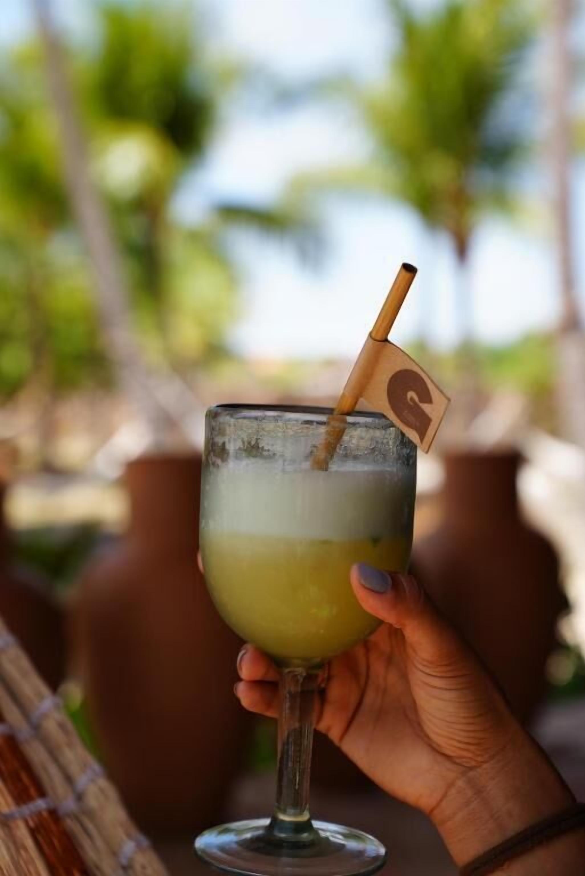 A hand holding a cocktail glass with a straw and a small flag, set against a blurred outdoor background.