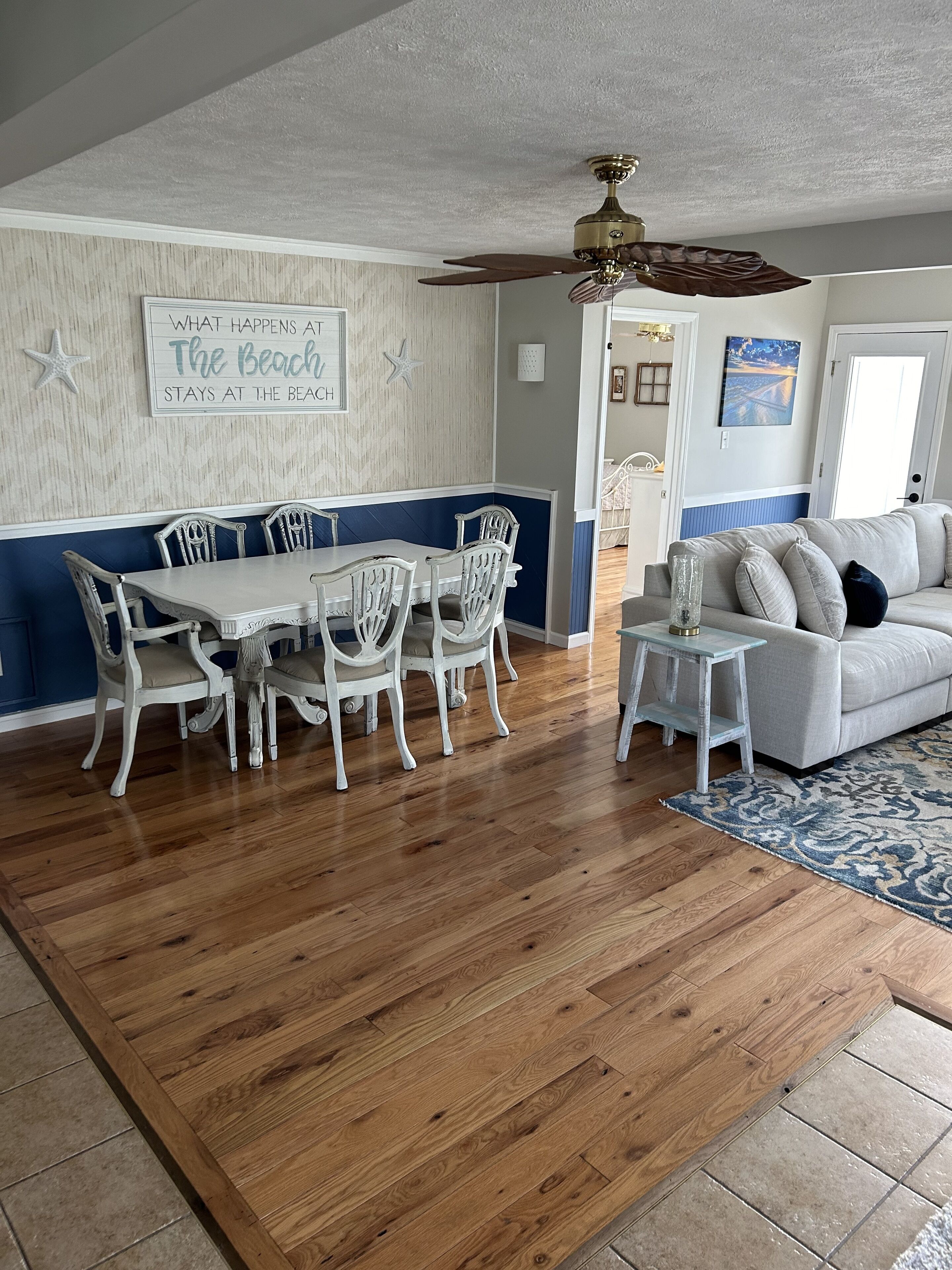 A dining area with a wooden table and white chairs, a ceiling fan, and a wall with a beach-themed sign.