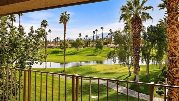 Vista da un balcone su un campo da golf con stagno, palme e cielo sereno.