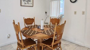 Un comedor con una mesa redonda de madera y seis sillas, un cuadro en la pared, un reloj de pared y una ventana con persianas.