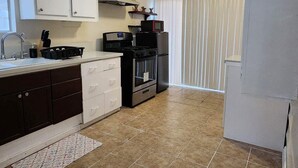 Cocina compacta con armarios blancos, estufa, microondas y refrigerador. Hay un tragaluz sobre el lavamanos y una ventana con persianas.