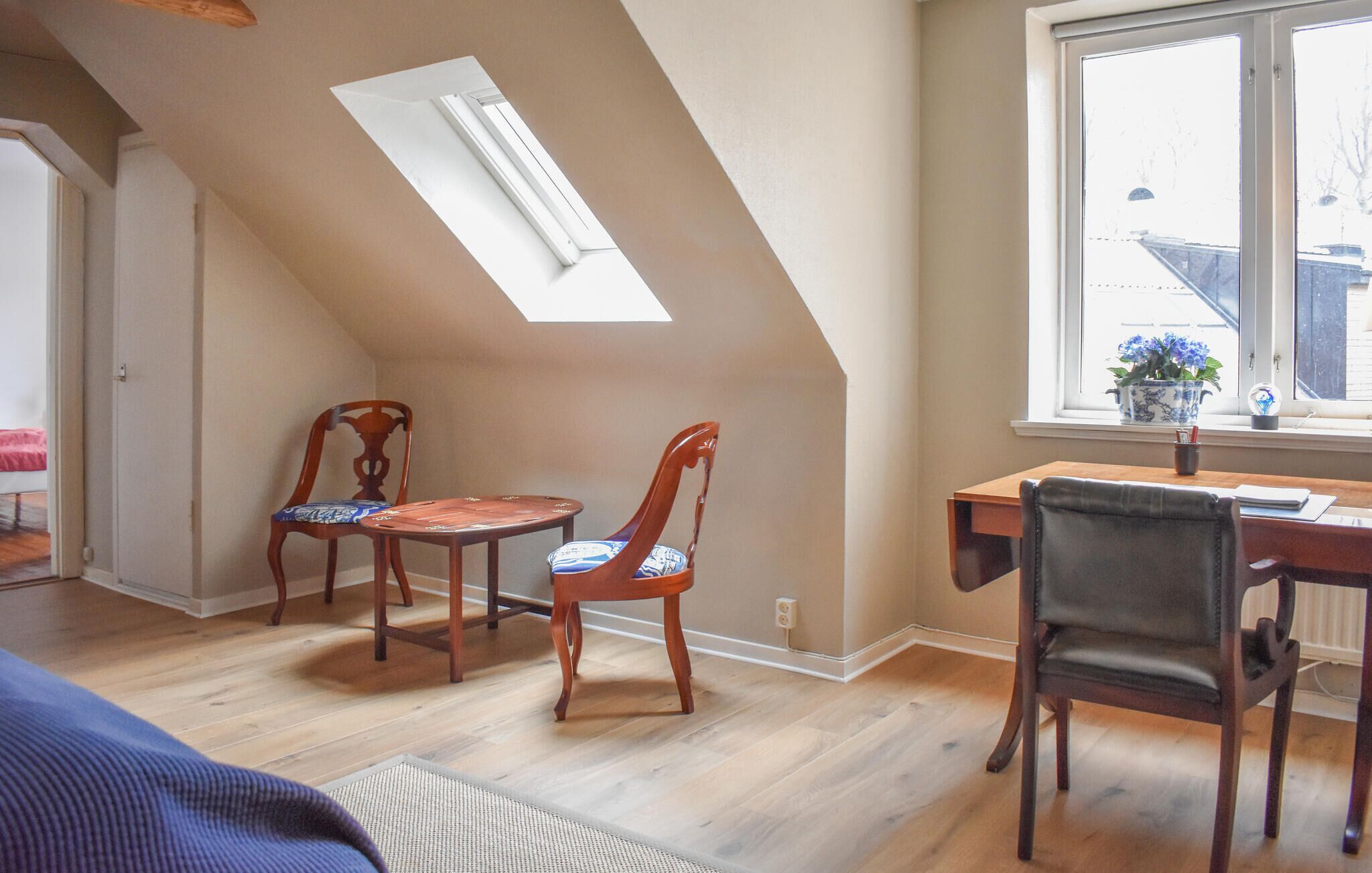 Une pièce avec un plafond en pente, deux chaises en bois dotées de coussins à motifs, une petite table en bois, un bureau avec une chaise et une fenêtre donnant sur les bâtiments environnants.